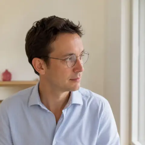 Portrait d’Antoine Dusserre, cheveux châtains courts et lunettes rondes, chemise bleu pâle, près d’une fenêtre