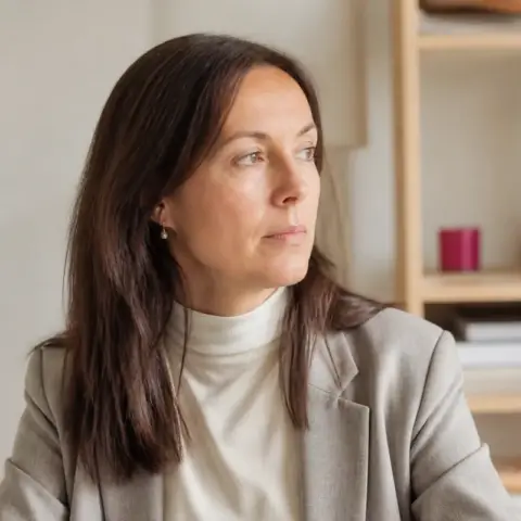 Portrait de Mathilde Marcenac, cheveux bruns mi-longs, blazer gris clair sur un col roulé écru, dans une pièce claire
